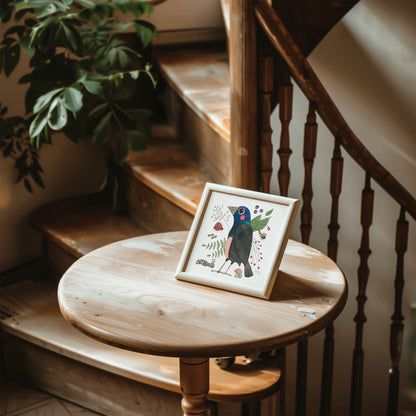 A framed art print featuring a colorful illustration of a common grackle surrounded by nature-inspired details, displayed on a round wooden table.