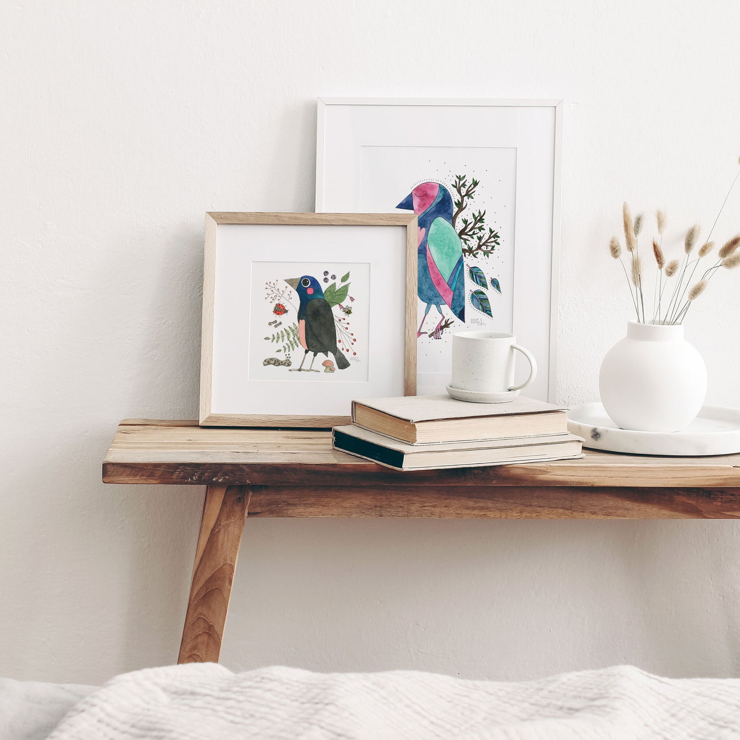 A framed art print featuring a colorful illustration of a common grackle surrounded by nature-inspired details, displayed next to a frame with another colorful bird, on a little wooden table.