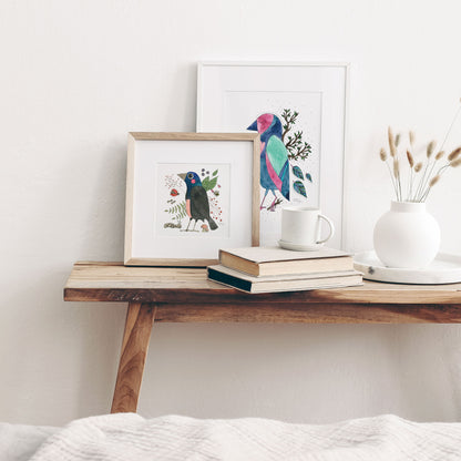 A framed art print featuring a colorful illustration of a common grackle surrounded by nature-inspired details, displayed next to a frame with another colorful bird, on a little wooden table.
