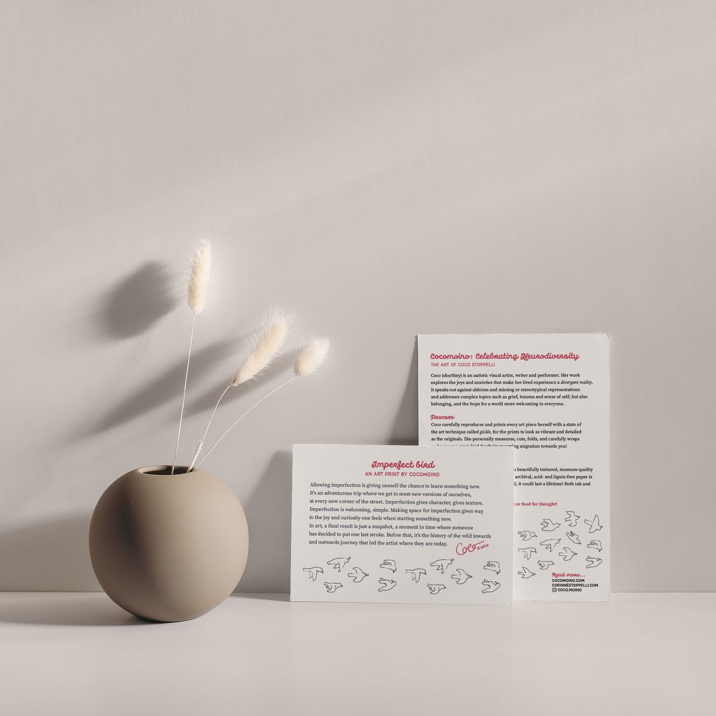 Two sheets of paper, a small one and a larger one, depicting the artwork's story and the production process, against a white wall, next to a vase