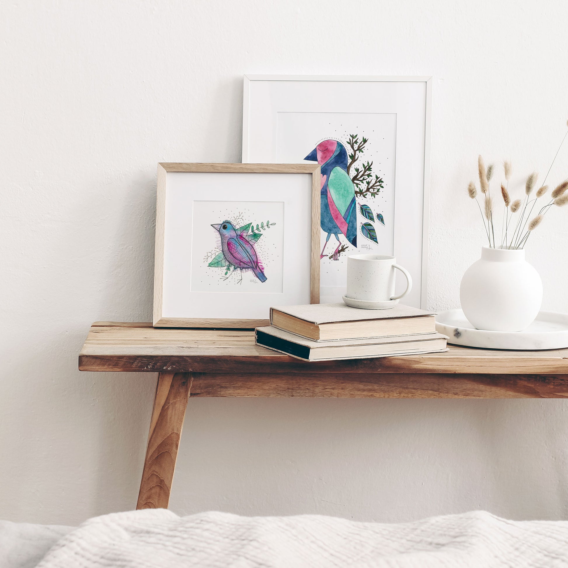 Two frames on a table against the wall, displaying two cute, colorful and whimsical bird watercolor artworks